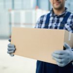 Close-up of moving company worker in gloves holding cardboard box and carrying it at container storage area